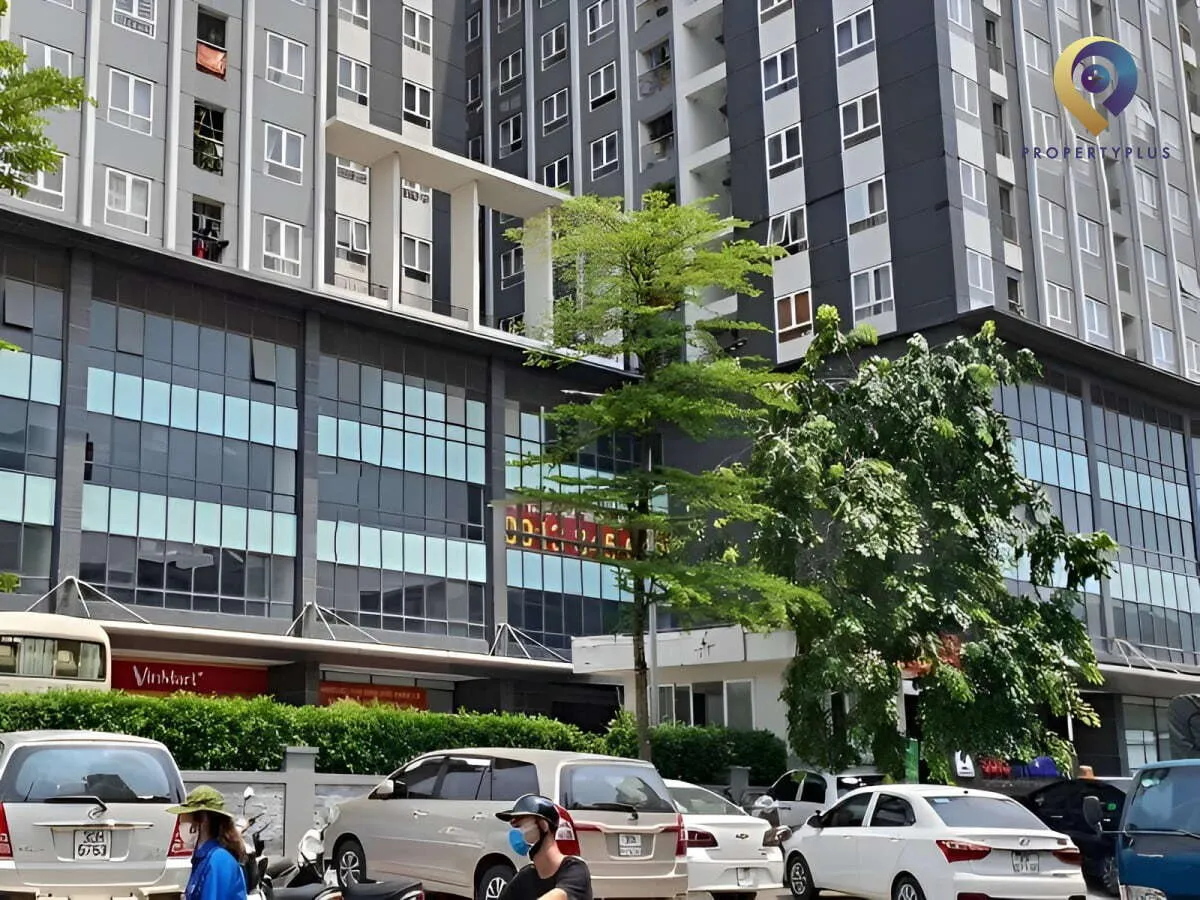 Khu vực phía trước tòa nhà UDIC-Riverside với bãi đỗ xe và cây xanh.